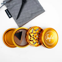 An open metallic gold herb grinder with three separated chambers, shown on a white background with a gray drawstring pouch and black scoop above it. The top lid has the brand name 'Jaglst' printed on it.