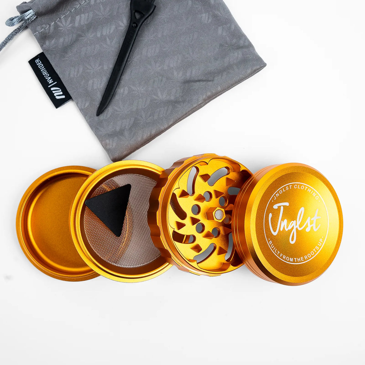 An open metallic gold herb grinder with three separated chambers, shown on a white background with a gray drawstring pouch and black scoop above it. The top lid has the brand name 'Jaglst' printed on it.