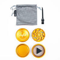 A metal herb grinder set with a gray drawstring pouch, black cleaning brush, and four gold-toned metal components on a white background. The top piece features the brand name 'Taglst' printed in white.