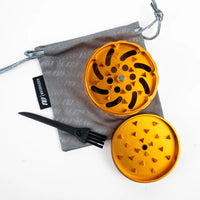 An orange metal herb grinder shown open with two circular pieces, a small black cleaning brush, and a gray drawstring pouch on a white background. A black fabric tag on the pouch reads 'M.