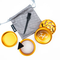 An open gold-colored metal grinder set with its lid, inner chamber, brush, and drawstring pouch on a white background. The brand name 'Jnglst' is clearly visible on the lid.