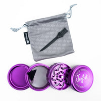 A purple metal herb grinder shown open with its inner components, alongside a gray drawstring pouch and a black spoon, on a white background. The pouch has a subtle cannabis leaf pattern, and the grinder lid features the brand name 'Jagdst'.
