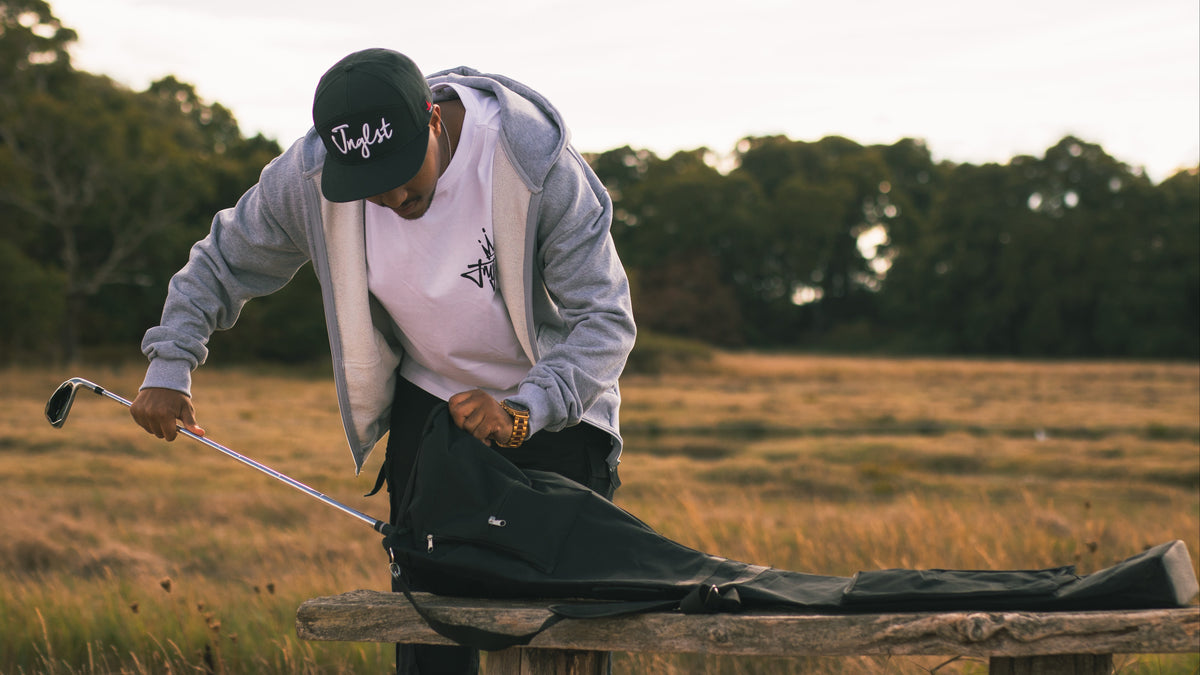 A man on a golf course bends over while putting a black golf bag on a wooden bench. He is wearing a gray zip-up hoodie, a white t-shirt, and a black baseball cap with the brand name TaylorMade visible on the front.