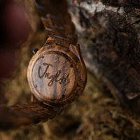 A close-up of a Jnglst wooden watch with a matching wood link band, resting against a textured tree bark background. The back of the watch case is engraved with the name 'Jnglst'.