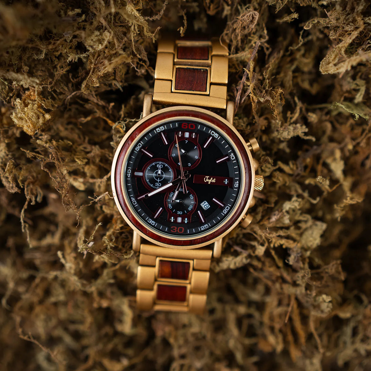 A gold-tone wristwatch with a black dial and red accents, featuring a metal link bracelet with dark wood inlays, resting on a bed of dried moss.