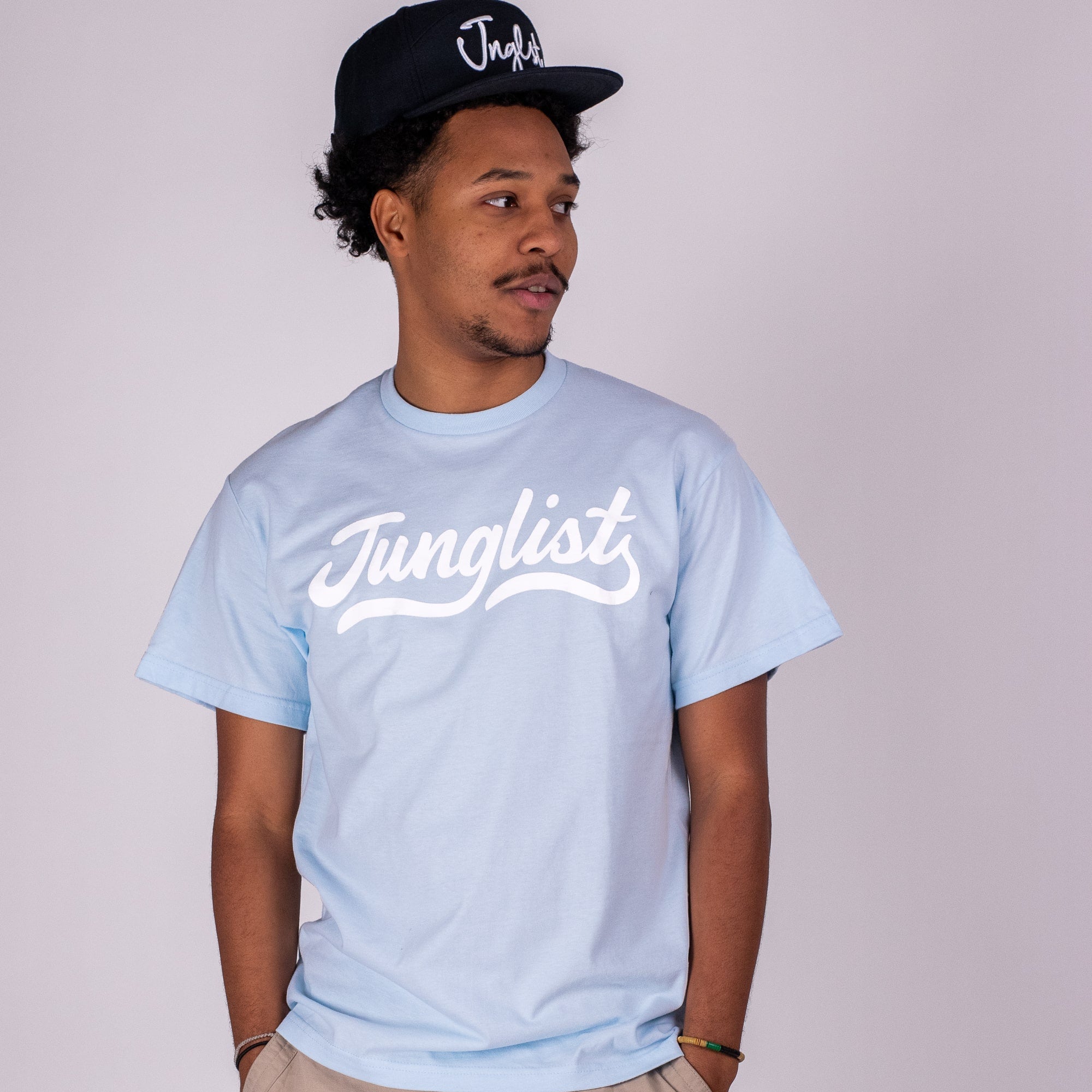 Man wearing a light blue 'Junglist' t-shirt and black cap on a plain background