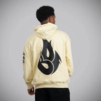 A person viewed from the back wearing a pale cream hoodie with a large black flame-like graphic on the back and black pants, standing against a light gray studio background.