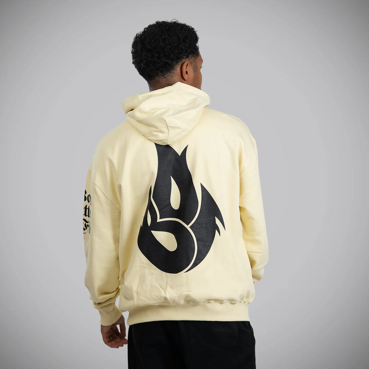 A person viewed from the back wearing a pale cream hoodie with a large black flame-like graphic on the back and black pants, standing against a light gray studio background.