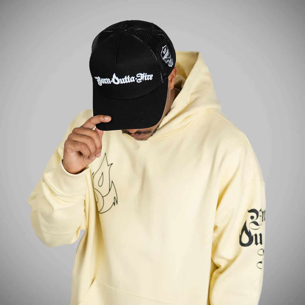 A person wearing a pale yellow hoodie and a black trucker cap with white Gothic-style lettering, posed against a neutral gray background.