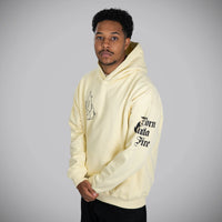 A man wearing a pale yellow hoodie with black graphic text on the sleeve and chest, standing against a neutral gray studio background.