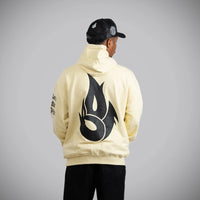 A person shown from the back wearing a cream-colored hoodie with a large black flame graphic on the back, black pants, and a black cap, photographed against a plain light gray studio background.