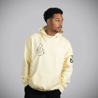 A man wearing a pale yellow hoodie with a black flame graphic on the left chest and a black graphic on the right sleeve, photographed against a solid gray background.