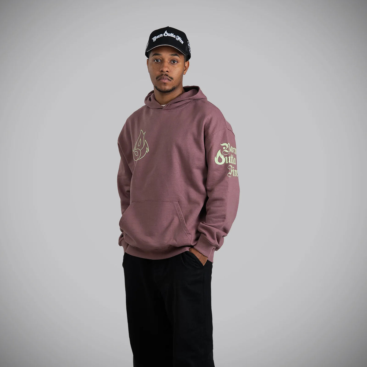 A man wearing a dusty rose hoodie with gold graphics on the chest and sleeve, paired with black pants and a black baseball cap, standing against a neutral gray background.