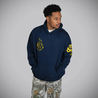 A man wearing a navy blue hoodie with yellow graphic prints on the chest and sleeve, paired with camouflage pants, standing against a neutral gray background.