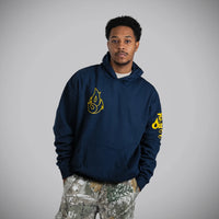 A man wearing a navy blue hoodie with yellow flame graphics on the chest and sleeve, paired with camouflage pants, standing against a solid gray background.