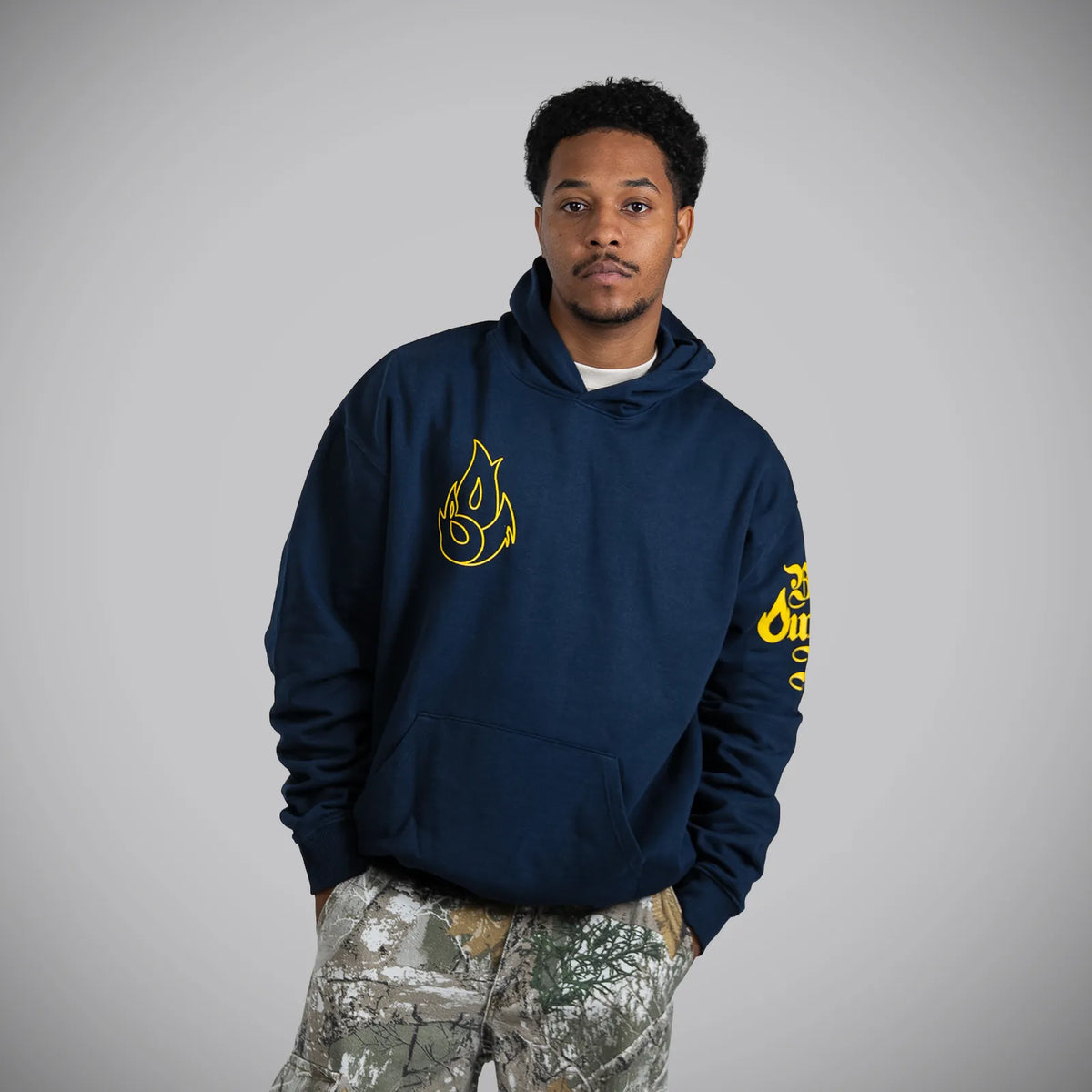 A man wearing a navy blue hoodie with yellow flame graphics on the chest and sleeve, paired with camouflage pants, standing against a solid gray background.