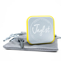A square metal jewelry box with rounded corners, featuring a silver glittery finish, a bright yellow border, and the brand name 'Jnglst' in white script. The box is displayed on top of a gray drawstring pouch with a black zipper pull on a clean white background.