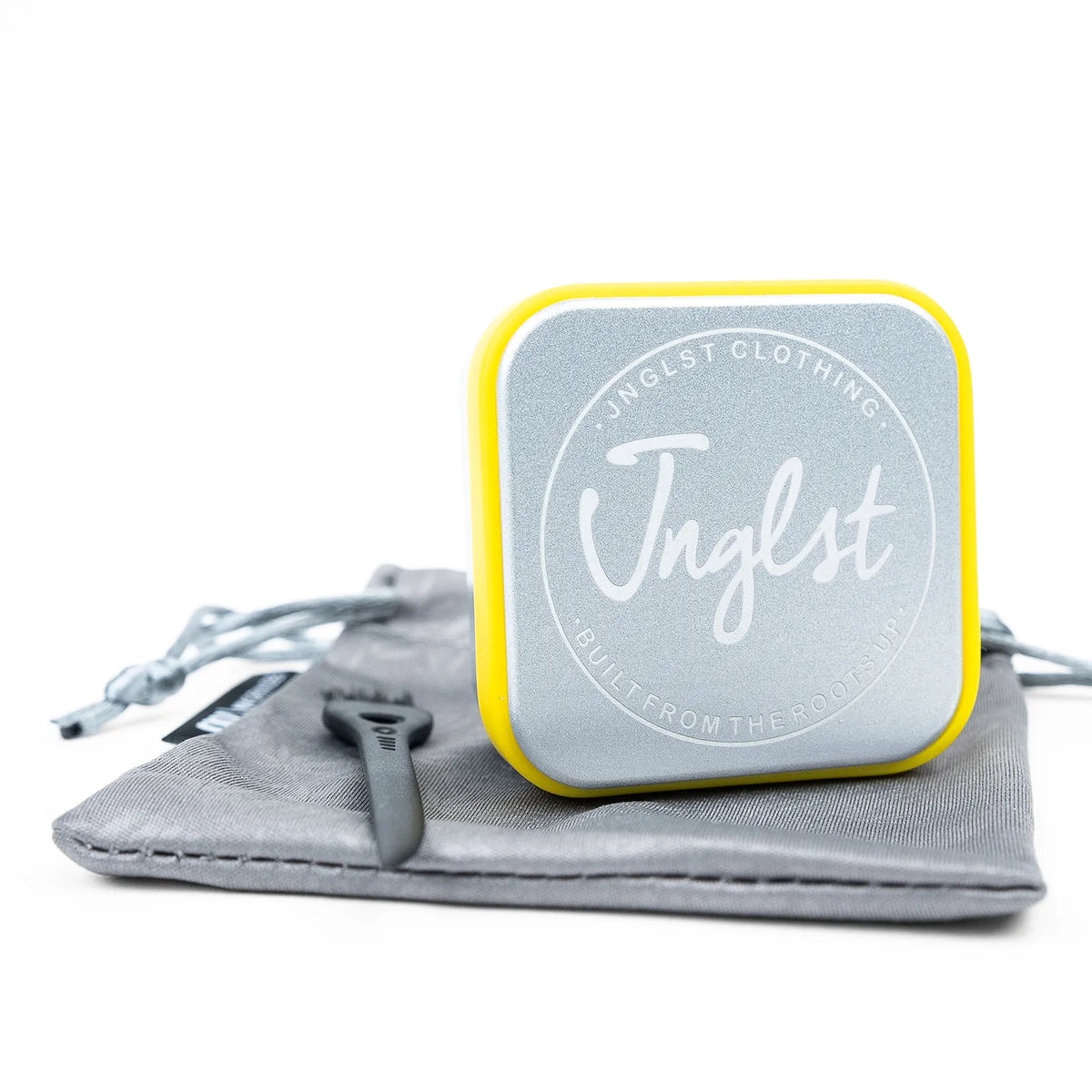 A square metal jewelry box with rounded corners, featuring a silver glittery finish, a bright yellow border, and the brand name 'Jnglst' in white script. The box is displayed on top of a gray drawstring pouch with a black zipper pull on a clean white background.