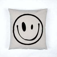 A square throw pillow in a light beige linen fabric featuring a large, hand-drawn black smiley face graphic in the center, set against a clean white background.