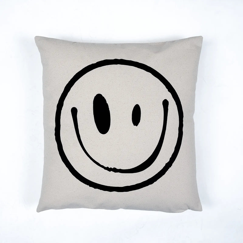 A square throw pillow in a light beige linen fabric featuring a large, hand-drawn black smiley face graphic in the center, set against a clean white background.