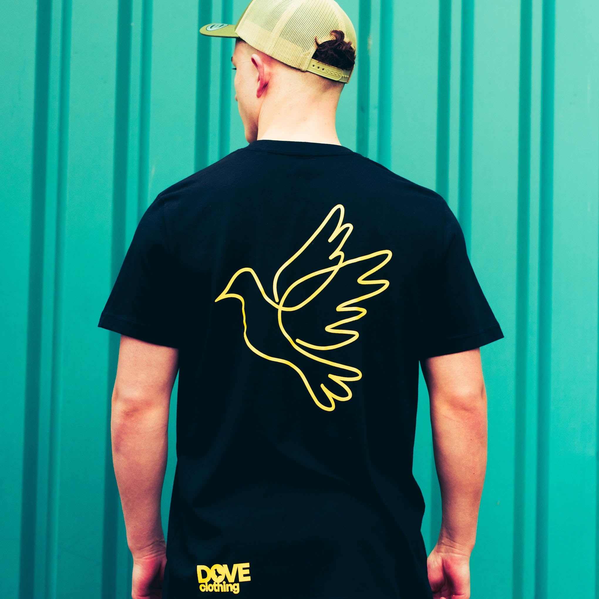 Person wearing a black t-shirt with a yellow dove design on the back, standing against a teal background.