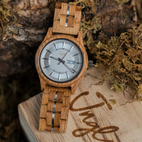 A wooden Jnglst watch with a natural wood grain strap and case, featuring a silver dial and a date window displaying 'SAT 26'. It is styled on a wooden block with dried moss and bark in the background.