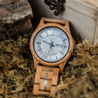A Junglist wristwatch with a light natural wood case and matching wood link bracelet, featuring a white dial with black markings and a day-date window. The watch is styled in moss next to a wooden box.