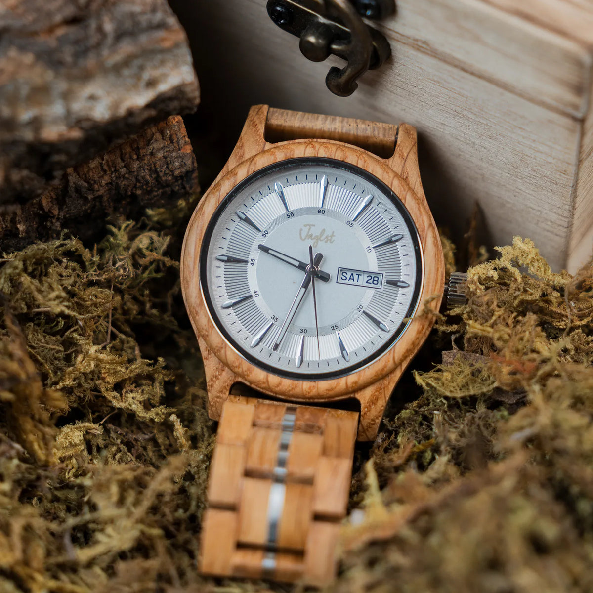 A Junglist wristwatch with a light natural wood case and matching wood link bracelet, featuring a white dial with black markings and a day-date window. The watch is styled in moss next to a wooden box.