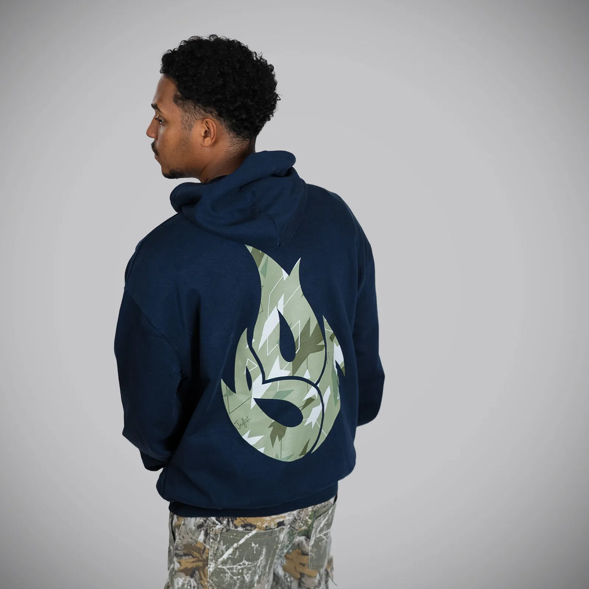 A man shown from the back wearing a navy blue hoodie with a large green camouflage flame graphic on the back, paired with camouflage pants, against a plain gray background.