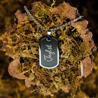 A silver-toned metal dog tag necklace with the name 'Jnglst' engraved in white script, resting on a textured bed of brown bark and green moss.