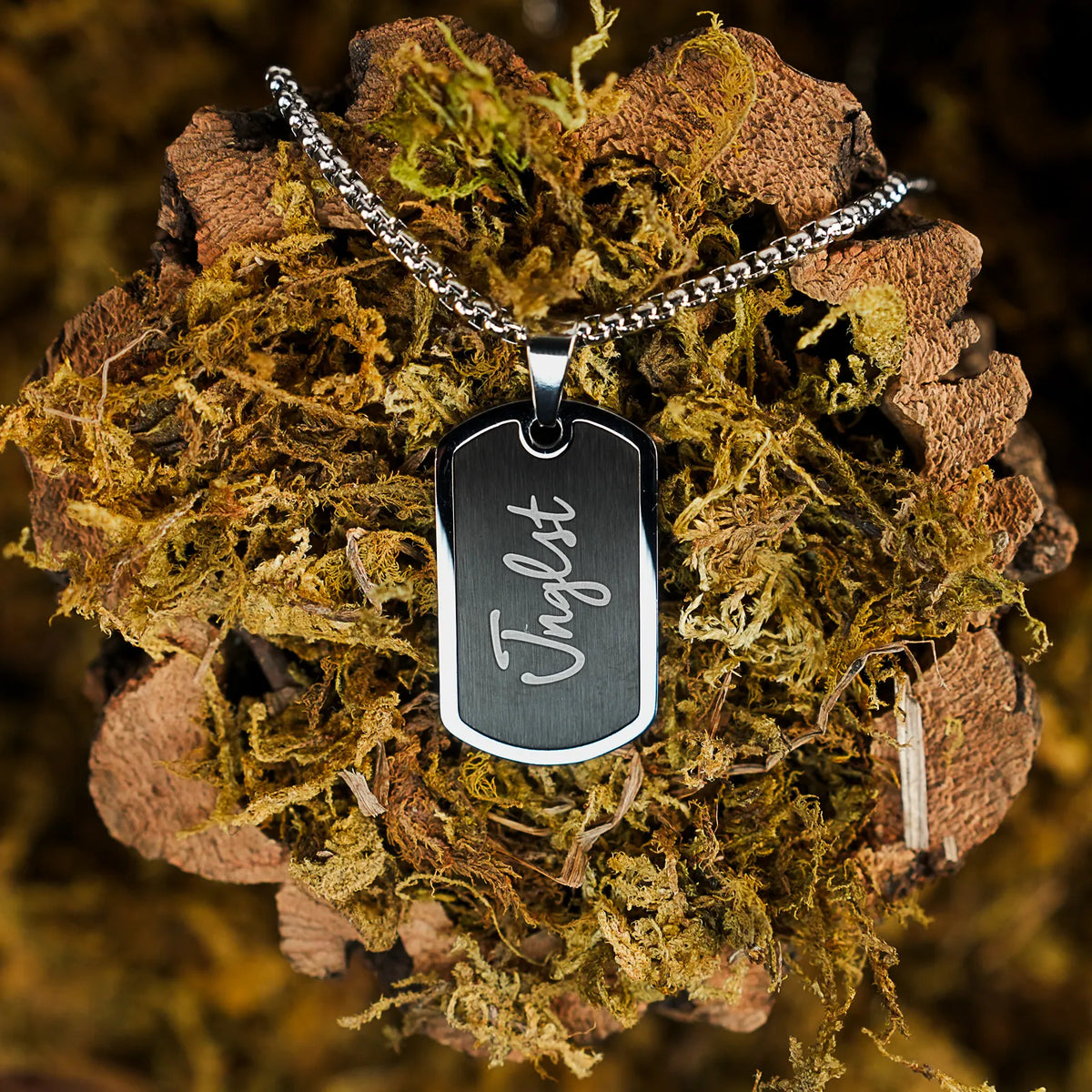A silver-toned metal dog tag necklace with the name 'Jnglst' engraved in white script, resting on a textured bed of brown bark and green moss.