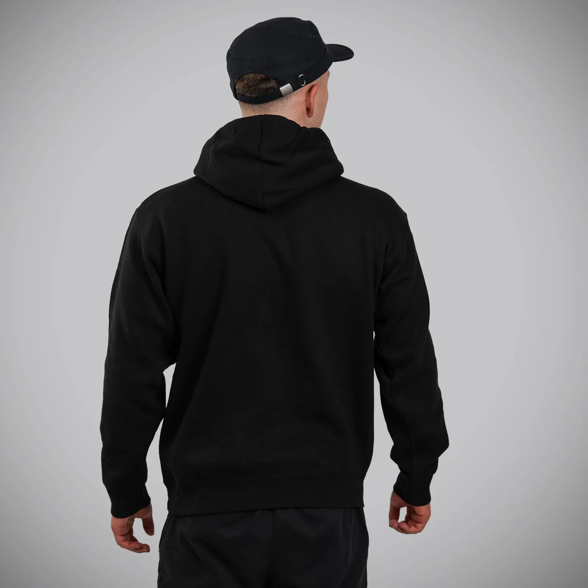 A man viewed from behind wearing a plain black pullover hoodie and a black baseball cap, standing against a light gray studio background.
