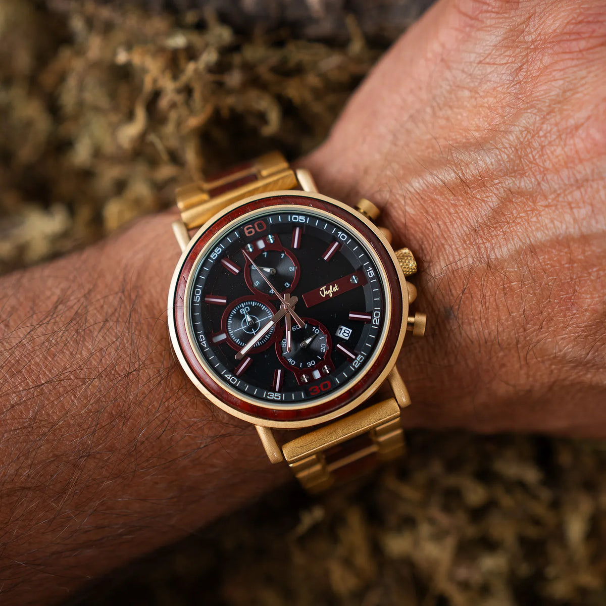 A gold-tone wristwatch with a black dial and red accents, shown worn on a person's wrist against a blurred natural background.