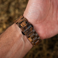 A close-up of a wooden link watch band worn on a person's wrist, showing the textured interlocking wood links in warm brown tones against a blurred natural background.