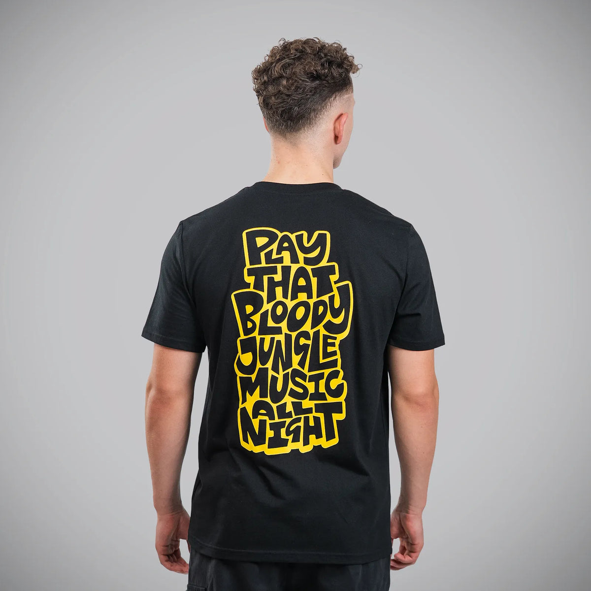 A man viewed from behind wearing a black t-shirt with a large yellow graphic text on the back reading 'PLAY THAT BLOODY JUNGLE MUSIC NIGHT'.