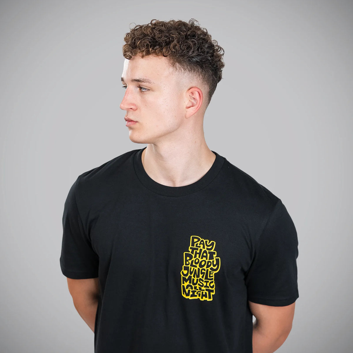 A man wearing a black crew-neck t-shirt with a yellow graphic text print on the left chest, photographed against a neutral gray background.