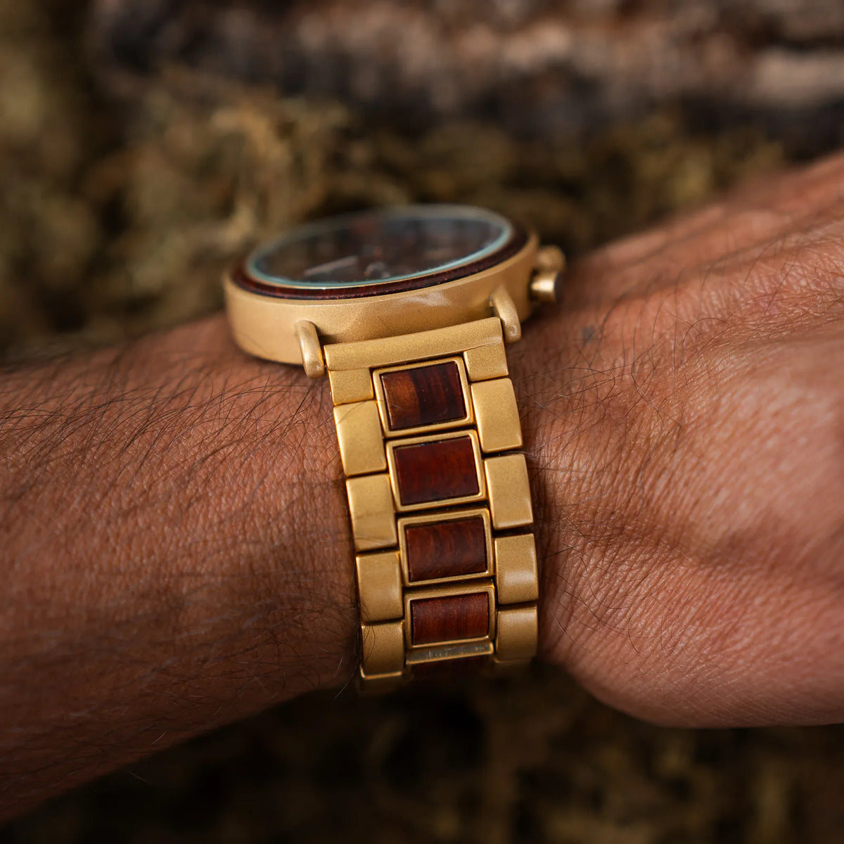 A close-up of a gold-tone wooden Jnglst watch with reddish-brown inlaid links, worn on a person's wrist against a blurred natural background.