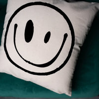 A square white throw pillow featuring a large black hand-drawn smiley face, resting on a dark teal velvet surface.