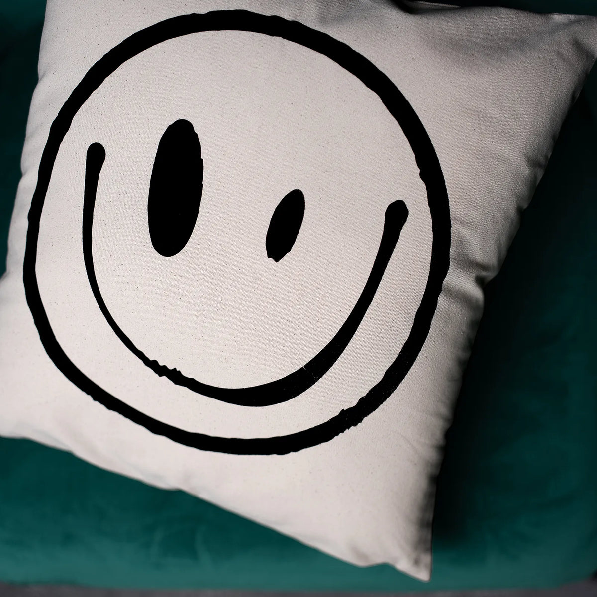 A square white throw pillow featuring a large black hand-drawn smiley face, resting on a dark teal velvet surface.