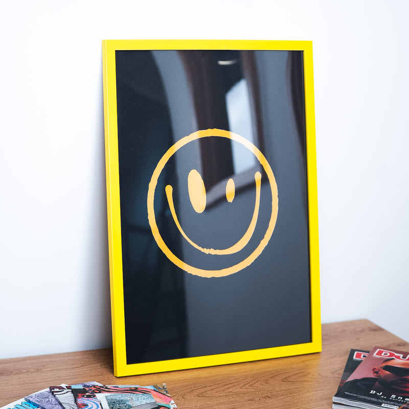 A framed art print featuring a yellow smiley face on a black background, housed in a bright yellow frame and leaning on a wooden table against a white wall.