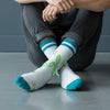 Person wearing white socks with green text and blue Jnglst accents on a gray floor.